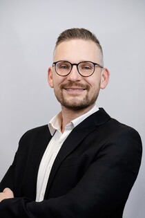 Porträtfoto eines Mannes mit Brille, Bart und schwarzem Sakko.